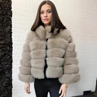 Eine Frau mit kastanienbraunem Haar steht mit dem Rücken zu einer weißen Steinmauer und trägt eine beigefarbene Daunenjacke aus Kunstpelz und eine schwarze Hose.