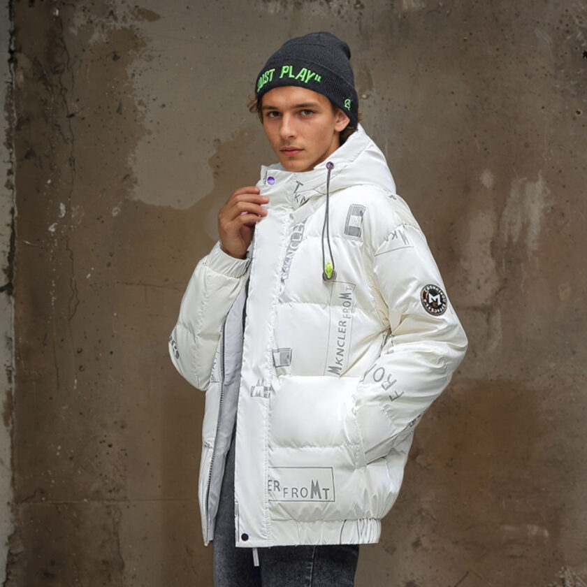 Junger Mann mit einer grauen Mütze mit grüner Schrift, der eine weiße gesteppte Daunenjacke mit aufgedruckter Schrift an der Kapuze und eine schwarze Hose trägt. Seine linke Hand steckt in der Tasche der Daunenjacke, die andere hält das Oberteil der Daunenjacke. Im Profil mit einer braunen Wand dahinter.