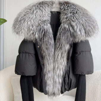 Schwarze, gesteppte Daunenjacke mit Strickärmeln und einem großen Kragen aus silbergrauem Fuchspelz. Sie ist auf einem Kleiderbügel vor einem weißen Sofa ausgestellt