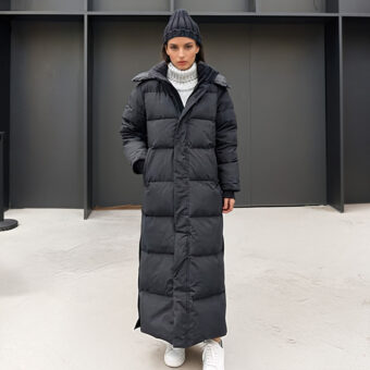 Stehende dunkelhaarige Frau mit weißen Schuhen, einer schwarzen Wollmütze und einer extra langen, schwarzen, gesteppten Daunenjacke. Ihre rechte Hand steckt in der Tasche der Daunenjacke. Im Hintergrund befindet sich eine schwarze Wand im Holzstil.