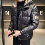 Wir sehen einen Mann in einer eleganten Hotellobby. Er ist schwarz gekleidet und trägt eine schwarze, glänzende Daunenjacke und die Kapuze ist mit einem beigen, karierten Stoff gefüttert.