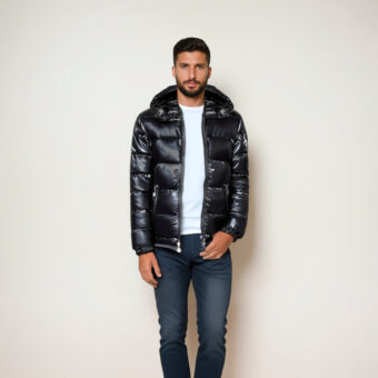 Ein unrasierter brauner Mann in blauen Jeans, einem weißen T-Shirt und einer glänzenden schwarzen Steppjacke mit Kapuze. Er steht vor einer beigen Wand.