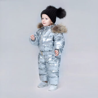 Stehendes Baby-Mädchen mit einer schwarzen Bommelmütze und einer glänzenden grauen Daunenjacke für Baby-Mädchen auf einem grauen Hintergrund.