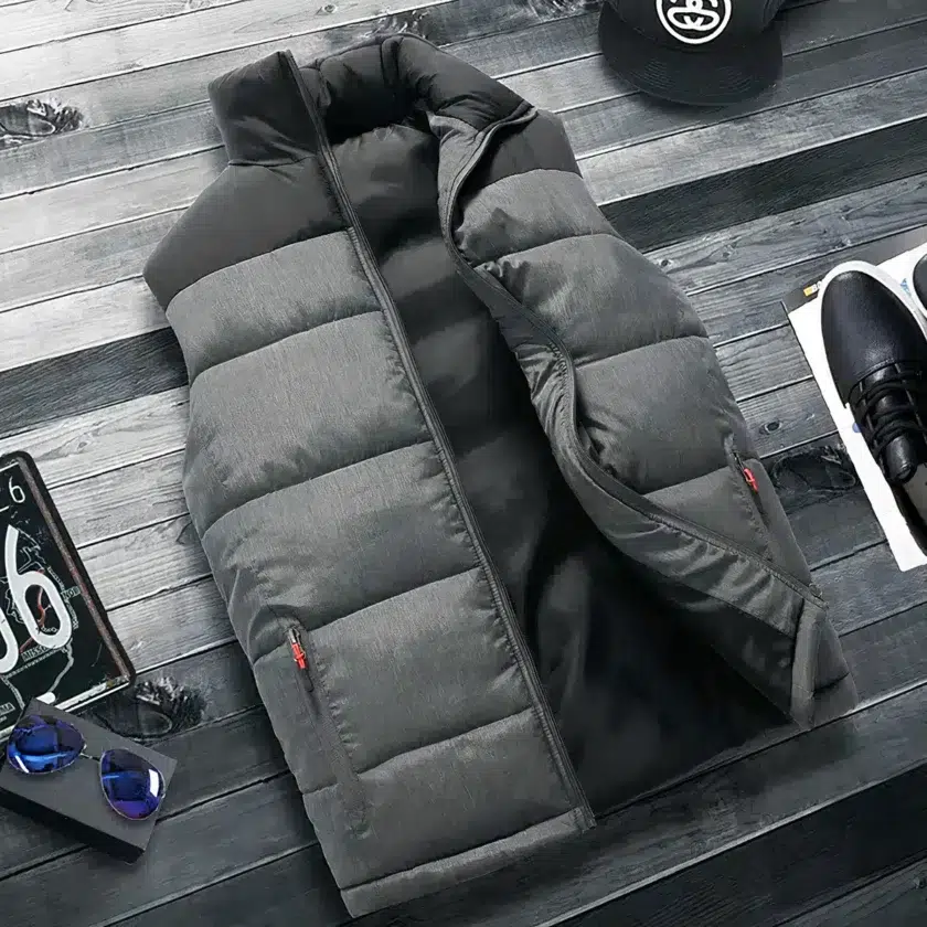 Graue und schwarze Daunenjacke auf dem Oberkörper, ärmellos gesteppt mit halboffenen Reißverschlusstaschen, auf dunkelgrauem Lattenparkett ausgebreitet. An den Seiten befinden sich eine schwarze Mütze, ein schwarzer Turnschuh und ein Stück des Nummernschildes.
