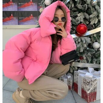Hockende Frau mit Sonnenbrille vor einem Weihnachtsbaum mit einer rosa Daunenjacke und Kapuze auf dem Kopf.
