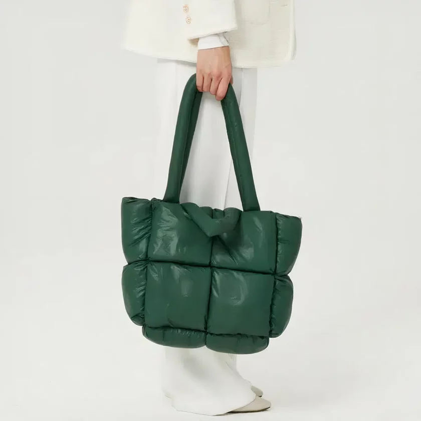 Frauenbeine im Profil mit weißer Hose auf weißem Hintergrund. Die Hand hält eine große, grüne, gesteppte Daunenjackentasche mit integriertem Schulterriemen.