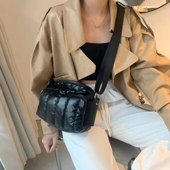 Sitzende Frau mit einer weißen Hose, einem schwarzen Oberteil und einer camelfarbenen Lederjacke, mit einer glänzenden, schwarzen, gesteppten Tasche über der Schulter