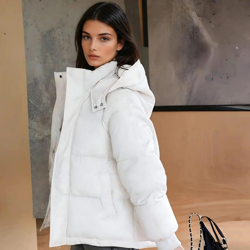 Braune Frau im Profil mit einer kurzen weißen Daunenjacke mit Kapuze. Eine Handtasche hinter ihr und eine beige-graue Wand.