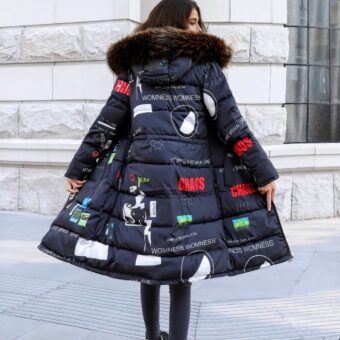 Eine dunkelhaarige Frau steht von hinten mit einer beigen Steinmauer vor sich, ihr Kopf ist nach rechts gedreht. Sie trägt eine schwarze Hose und eine lange, wendbare Daunenjacke in Marineblau mit weißen, roten und grünen Mustern und Aufschriften. Die Kapuze ist am Rand mit Pelz besetzt.