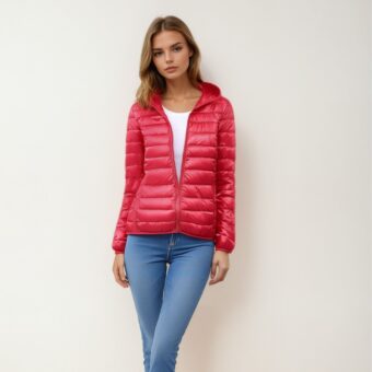 Blonde Frau steht mit dem Rücken zu einer beigen Wand und trägt eine blaue Röhrenjeans, ein weißes Oberteil und darüber eine leichte, warme, rote Daunenjacke, die mit einem Reißverschluss zu einem Drittel geschlossen ist, so dass das weiße Oberteil zu sehen ist.