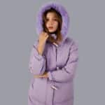 Frau in einer violetten Daunenjacke mit einer pelzigen Kapuze auf dem Kopf, auf grauem Hintergrund.