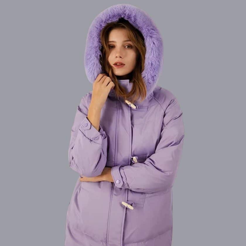 Frau in einer violetten Daunenjacke mit einer pelzigen Kapuze auf dem Kopf, auf grauem Hintergrund.