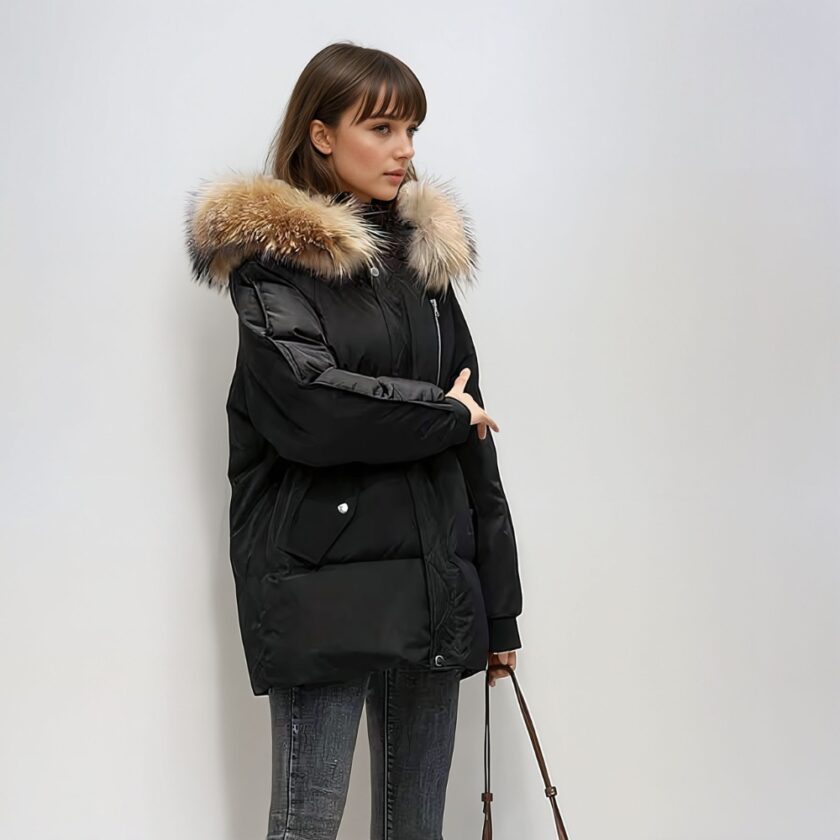 Braune Frau mit Pony, vor einer weiß/grauen Wand, trägt eine schwarze Daunenjacke mit Kapuze und braunem, halblangem, gestepptem Pelz. Sie hält den Riemen einer Handtasche in ihrer linken Hand.