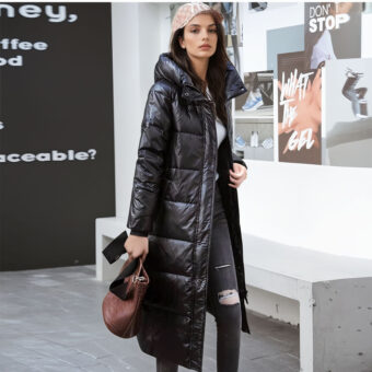 Stehende Frau mit einer beschrifteten Wand im Hintergrund auf der rechten Seite und einer schwarzen Wand auf der linken Seite mit weißer Beschriftung. Eine Bank aus grauem Stein hinter der Frau. Sie trägt eine schwarze Dlim-Hose mit einem Loch am linken Knie, eine beigefarbene Kappe, eine lange schwarze Kuscheldecke mit Kapuze und eine Handtasche in ihrer rechten Hand.