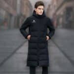 Schwarze, lange, wasserdichte Unisex-Fußballjacke, die von einem jungen Mann mit braunen Haaren getragen wird. Er scheint sich auf einer Straße zu befinden.
