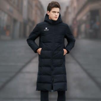 Schwarze, lange, wasserdichte Unisex-Fußballjacke, die von einem jungen Mann mit braunen Haaren getragen wird. Er scheint sich auf einer Straße zu befinden.