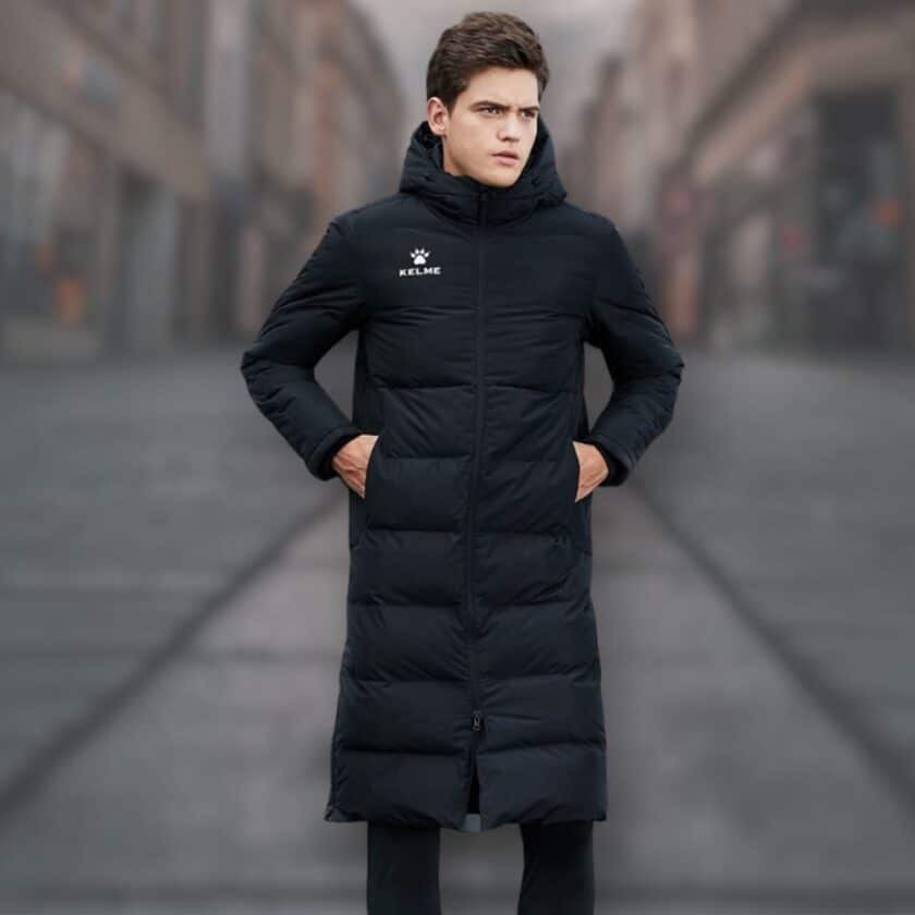 Schwarze, lange, wasserdichte Unisex-Fußballjacke, die von einem jungen Mann mit braunen Haaren getragen wird. Er scheint sich auf einer Straße zu befinden.