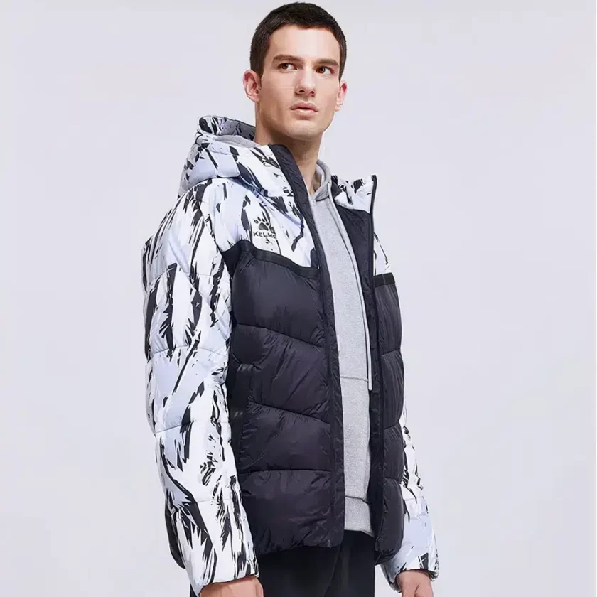 Stehender brauner Mann auf grauem Hintergrund, der eine kurze, offene Fußballjacke trägt, die einen grauen Pullover sichtbar macht. Die Daunenjacke ist am unteren Teil des Oberkörpers einfarbig schwarz gesteppt und am oberen Teil der Schultern und an den Ärmeln schwarz-weiß getigert.