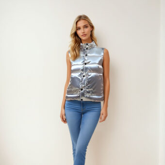 Blonde Frau, die an einer beigen Wand steht und eine blaue Röhrenjeans und eine silberne ärmellose Daunenjacke trägt.