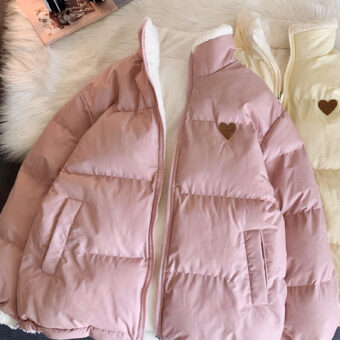 Wir sehen zwei Daunenjacken: eine hellrosa und eine beige. Beide haben ein kleines gesticktes Herz auf der Brust. Sie sind beidseitig tragbar und die andere Seite ist aus Fleece mit einem Teddybär-Motiv auf der Brust. Sie werden auf einem Teppich aus Schafsfellimitat neben einem Kopfhörer präsentiert. Das Foto wurde von oben aufgenommen.