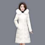 Brünette asiatische Frau, die eine lange, gesteppte weiße Daunenjacke mit Pelzkragen trägt. Ihre rechte Hand steckt in der Tasche der Daunenjacke. Auf grauem Hintergrund.