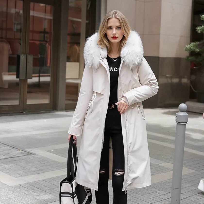 Braune Frau mit halblangem Haar, die auf einer Straße mit einem Gebäude im Hintergrund steht. Sie trägt eine enge schwarze Hose, ein schwarzes Oberteil und eine weiße Daunenjacke in Übergröße mit einer gleichfarbigen Kapuze aus Kunstpelz.