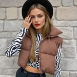 Eine Frau mit langen blonden Haaren, die eine schwarze Hose, ein kurzes, langärmeliges Oberteil mit einem schwarz-weißen Zebramuster, eine braune, gesteppte, ärmellose Daunenjacke und einen schwarzen Hut trägt. Sie steht mit dem Rücken zu einer beigen Steinmauer.