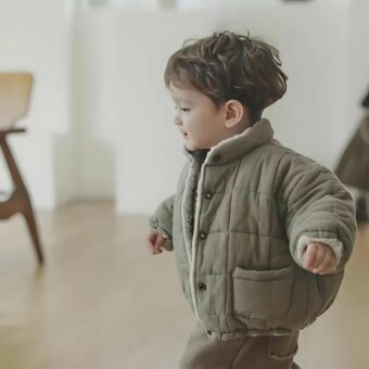Kleiner Junge, der die dicke, warme, fleecegefütterte Baby Daunenjacke in der khakifarbenen Version trägt. Der kleine Junge trägt sie in seinem Haus, in seinem Esszimmer, wie es scheint.