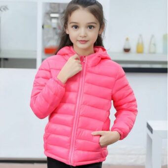 Kleines Mädchen, das eine dünne Daunenjacke mit Kapuze für Kinder trägt und sich in einem Raum befindet, der eine Küche zu sein scheint.
