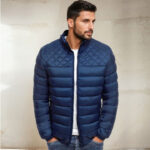 Brauner Mann mit kurzem Bart, der vor einer beigen Wand steht und Jeans, ein weißes T-Shirt und eine leichte, dünne, taillierte Daunenjacke in Marineblau trägt. Seine Hände stecken in den Taschen der blauen Jeans.