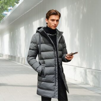 Halblange Daunenjacke für Männer in grauer Ausführung, die von einem jungen Mann mit braunen Haaren getragen wird, der sein Handy in der linken Hand hält. Er steht auf der Straße.