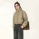 Eine Frau asiatischen Typs mit zusammengebundenen Haaren und einer Brille im Stehen. Sie trägt eine schwarze Hose und eine kurze khakifarbene Daunenjacke aus Kunstleder. Ihre Hände befinden sich in den Taschen der Hose. Über ihrer linken Schulter trägt sie eine schwarz-braune Handtasche. Auf beigem Hintergrund.