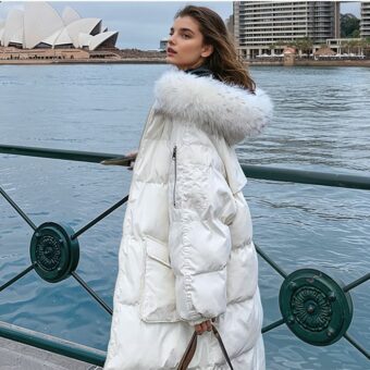 Eine Frau mit braunem Haar steht links von einem grünen Metallzaun. Rechts von diesem Zaun befindet sich eine flussähnliche Wasserfläche und im Hintergrund sind Gebäude zu sehen. Die Frau trägt eine lange weiße Daunenjacke mit weißer Pelzkapuze. In ihrer linken Hand hält sie einen Taschenriemen. Sie ist im Profil zu sehen.