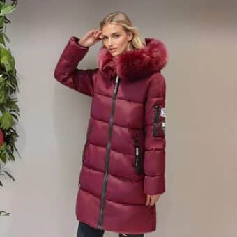 Blonde Frau im Profil neben einer hellbraunen Wand, die eine warme, halblange, bordeauxfarbene Daunenjacke mit passendem Pelz an der Kapuze trägt. Seitentaschen. Sie hält ihre rechte Hand neben ihrer Schläfe. Auf der linken Seite ist eine grüne Pflanze zu sehen.