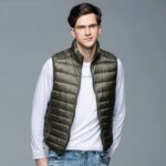 Mann vor grauem Hintergrund, Hände in den Gesäßtaschen in hellblauen Jeans, trägt eine ärmellose, khakifarbene Daunenjacke über einem weißen T-Shirt mit langen Ärmeln.