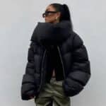 Frau in einer kurzen schwarzen Daunenjacke mit einem Oversize-Rollkragen, der mit einem silbernen Reißverschluss geschlossen wird, langen, weiten Ärmeln, die von vorne mit einer eng anliegenden schwarzen Hose posiert.