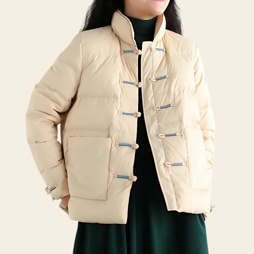 Elegante Tang Daunenjacke für Damen in beige mit raffinierten Froschverschlüssen, geräumigen Taschen und Stehkragen. Eine Frau trägt sie über einem schwarzen Kleid. Sie hat ihre Hände auf dem Rücken.