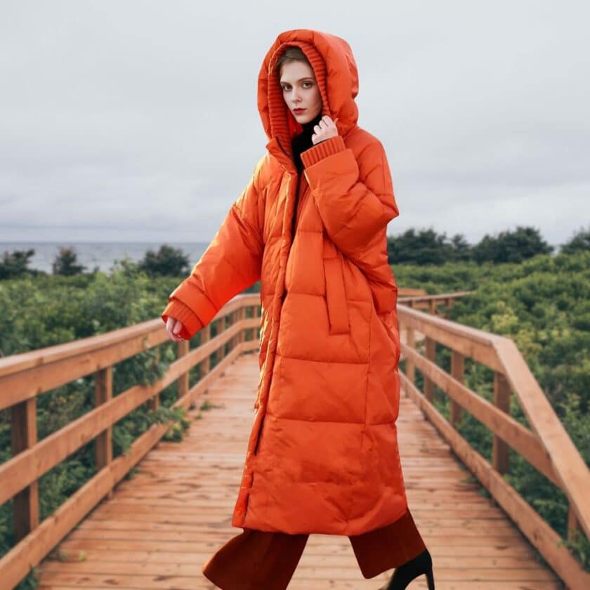 Orangefarbene Daunenjacke für Damen in großen Größen mit geprägter Kapuze und Entendaunen. Ansicht einer Frau, die diese lange Daunenjacke trägt, die ideal ist, um mollige Silhouetten im Winter mit einem Hauch von Fantasie und Wärme zu betonen.